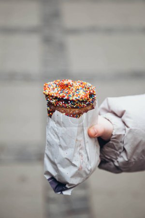Tourist holds Trdelnik decorated with colorful sprinkles against street. Grilled rolled dough topped with sugar. Traditional Czech hot sweet pastry.