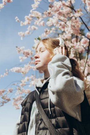 Une adolescente écoute de la musique sur écouteurs dans le contexte de fleurs de sakura un jour de printemps ensoleillé. Concept de liberté et de bonheur. Printemps..