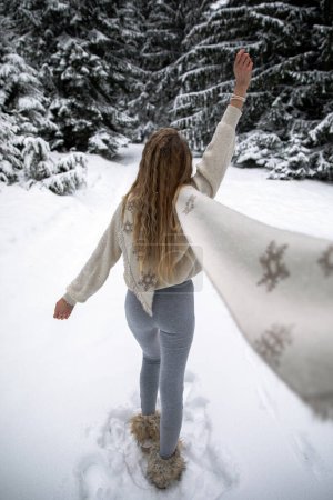 Téléchargez les photos : A young blonde girl with a long white scarf and a wool blouse walks through the snowy forest. Winter holiday concept. Back view. - en image libre de droit