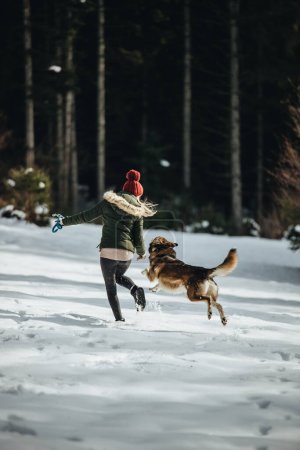 Foto de Young happy female and her dog in having fun in forest in winter season. - Imagen libre de derechos