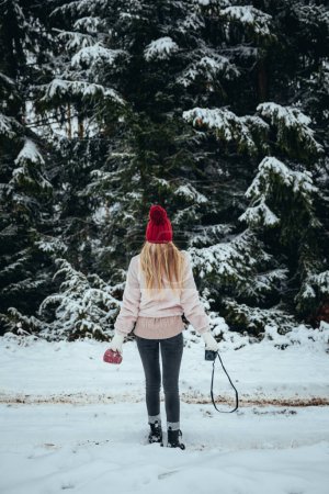 Foto de Back view of blonde woman with camera and red cup in hands standing on the snow covered forest - Imagen libre de derechos