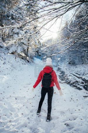 Foto de Backpacker young woman wearing red jacked and white hat. Enjoying the sunny weather outdoors after first snow in the woods. - Imagen libre de derechos
