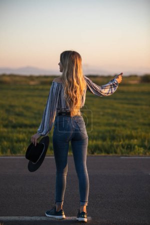Téléchargez les photos : Young attractive blonde woman hitchhiking along a road at sunrise. Freedom, holiday concept. - en image libre de droit
