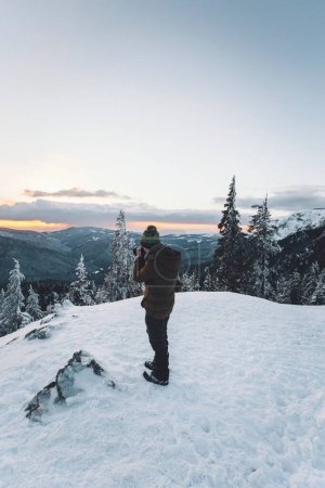 Foto de Fotógrafa profesional tomando fotos con su cámara de un árbol cubierto de nieve y montañas al atardecer - Imagen libre de derechos