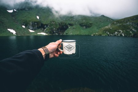 Téléchargez les photos : Young male traveler holding a mug with coffee on the mountain lake background . Adventure, lifestyle concept. - en image libre de droit