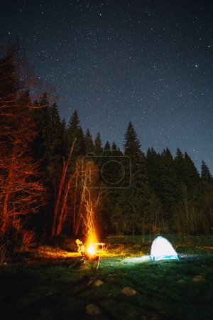 Foto de Tienda iluminada y fogata bajo el hermoso cielo nocturno lleno de estrellas en el bosque salvaje - Imagen libre de derechos