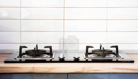 Téléchargez les photos : Vue de face de la cuisinière à gaz en verre trempé contemporain avec deux brûleurs avec bouton d'allumage automatique sur le comptoir en bois, la casserole en fonte supporte le capot du ventilateur et le four construit dans compact - en image libre de droit