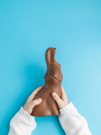 Photo for Hands of Child holding chocolate Easter bunny with ears bitten off on blue background, family concept. Vertical, top view - Royalty Free Image