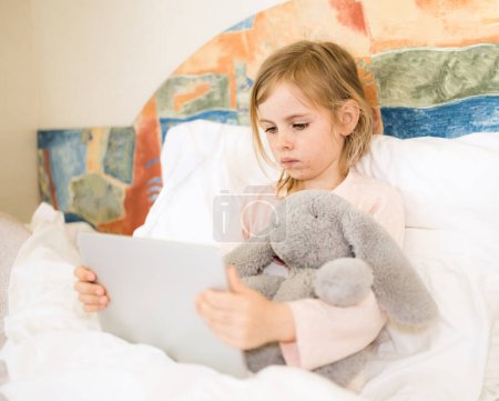 Sick child with red pimples sitting in bed hugging fluffy toy. Little girl looking at tablet. Chickenpox, varicella virus or vesicular rash on child's body and face