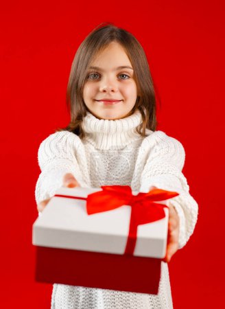 Photo for Child with arms extended forward holding gift box with red ribbon and bow. Little girl looking at camera. Kid wearing white sweater on red background, focus on foreground - Royalty Free Image