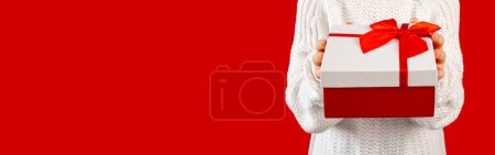 Photo for Unrecognisable child holding gift box with red ribbon and bow. Little girl wearing white sweater on red background, copy space - Royalty Free Image