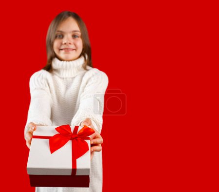 Photo for Happy child with arms extended forward giving or receiving gift box wrapped with red bow. Little girl smiling and looking at camera. Kid wearing white sweater on red background, focus on foreground - Royalty Free Image