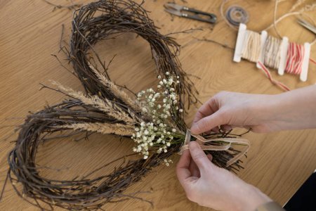 Hands tying bow on wicker wreath of birch branches on wooden background. Wreath weaving, handmade decoration