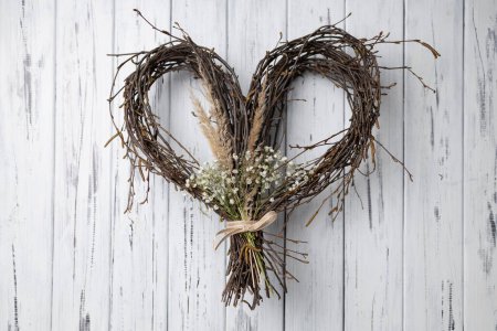 Crafted Wicker heart shaped wreath of birch branches hanging on white aged wooden background. Handmade DIY decoration