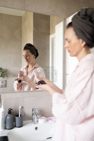 Frau im Turban quetscht Kosmetikcreme im Badezimmer aus. Konzept der Morgenpflege Routine, Anti-Age-Pflege. Fokus auf Reflexion im Spiegel