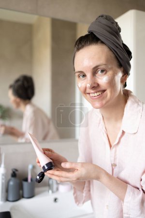 Mujer sonriente en turbante después de lavar el cabello mirando a la cámara y exprimiendo la crema cosmética en el baño. Concepto de rutina matutina de cuidado de la piel, cuidado anti-edad. Hidratación. Mancha de crema en la cara