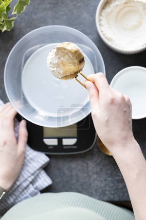 Téléchargez les photos : Main de femme tenant cuillère à mesurer avec de la farine. Balance, eau et farine sur le comptoir de la cuisine. Peser la farine pour la cuisson, la fabrication et l'alimentation du levain à la maison. Vue du dessus - en image libre de droit
