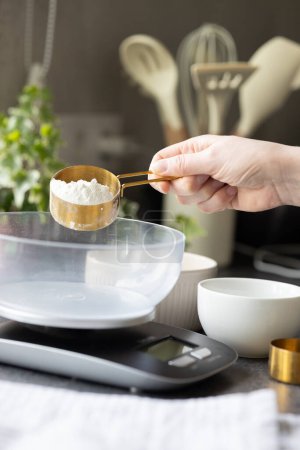 Téléchargez les photos : Main de femme tenant cuillère à mesurer avec de la farine. Peser la farine pour la cuisson, la fabrication, l'alimentation du levain à la maison. Balance, eau et farine sur le comptoir de la cuisine - en image libre de droit