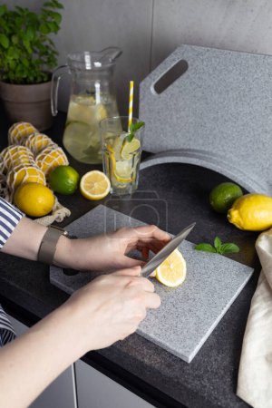hands of woman cutting lemon on stone cutting board. Lemonade preparation. Whole lemons and limes in string bag, carafe and pot with herbs on kitchen counter