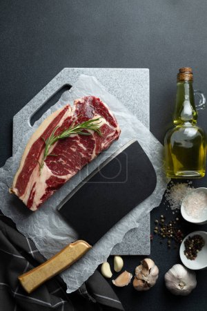 Fresh raw beef steak garnished with rosemary lies on cutting board. Salt, peppercorns, bottle of olive oil, garlic cloves, and meat cleaver with wooden handle on kitchen counter
