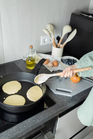 Child hand holding ladle above frying pan with pancakes on stove. Cutting board, blueberries in bowl, eggs, olive oil in bottle on kitchen counter. Preparation of healthy meal, family time, copy space
