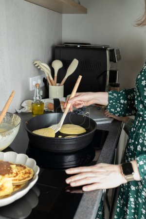 Woman hand holding spatula with pancakes in frying pan on stove. Kitchen utensils, olive oil in bottle on kitchen counter. Cooking at home, family time, copy space