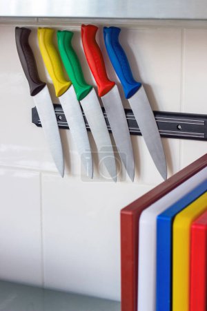 Photo for Five kitchen knives with colorful handles hang on a magnetic holder. Multicolored kitchen cutting boards. Professional kitchen equipment for chefs.Food safety. HACCP. Place for text - Royalty Free Image