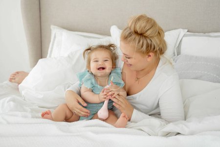 Photo for Laughing Mom and baby girl are lying in bed, enjoying time together. Family time concept. Funny playing with toy. - Royalty Free Image