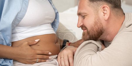 Téléchargez les photos : Beau homme écoute et regarde ventre enceinte de sa belle femme noire et sourit. Mariage multiracial. Parentalité. Gros plan - en image libre de droit