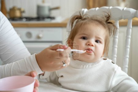 Photo for Baby girl eating blend mashed food sitting, on high chair, mother feeding child, hand with spoon for vegetable lunch, baby weaning, first solid food for kid. Healthy eating for baby humans - Royalty Free Image