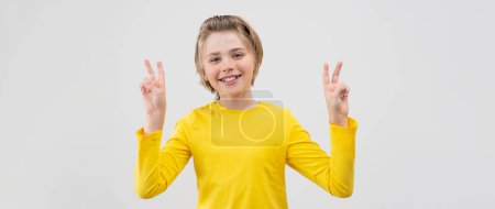Foto de Feliz adolescente sonriente mostrando signo de victoria, paz sobre fondo blanco. Niño rubio con el pelo largo y camiseta amarilla agitando las manos Emocionado. Copiar espacio - Imagen libre de derechos