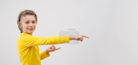Astonished Emotional Teenager boy happily Joy Showing To Right Over White Background. Copy Space For Advertisement. Excited Blond Kid With Long Hair And Yellow T-Shirt