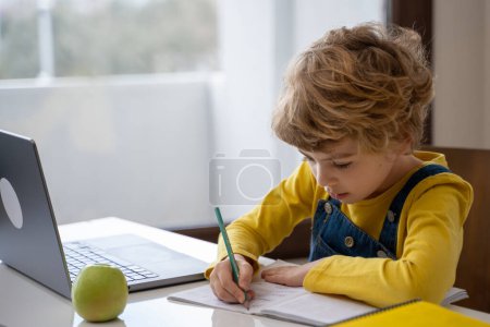 Foto de Caucasian child schoolboy or girl studying at home using laptop remote education. Doing homework, writing exercise book. Copy space - Imagen libre de derechos