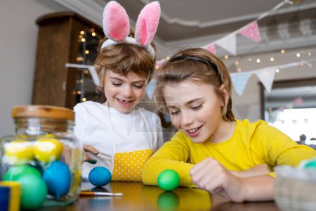 Téléchargez les photos : Easter Family traditions. Two caucasian happy children with bunny ears dye and decorate eggs with paints for holidays playing together. Kids having fun. - en image libre de droit