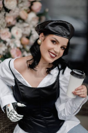 Foto de Beauty disabled french styled Woman with bionic prosthetic arm, artificial hand drink coffee outside in spring flower interior cafe. Vertical. Women diversity - Imagen libre de derechos