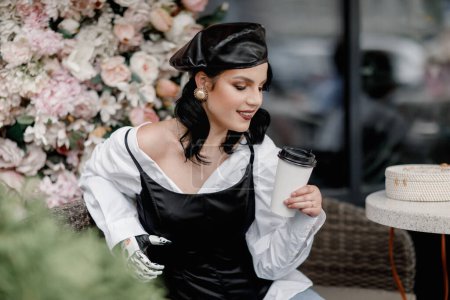 Foto de Beauty disabled french styled Woman with bionic prosthetic arm, artificial hand drink coffee outside in spring flower interior cafe. Vertical. Women diversity - Imagen libre de derechos
