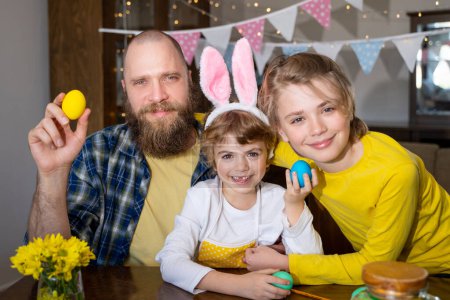 Photo for Easter Family traditions. Father and two caucasian happy children with bunny ears dye and decorate eggs with paints for holidays while sitting together at home table. Kids embrace and smile in cozy. - Royalty Free Image