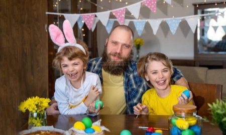 Téléchargez les photos : Easter Family traditions. Father and two caucasian happy smiling children with bunny ears dye and decorate eggs with paints for holidays while sitting together at home table. Dad embrace and smile in - en image libre de droit