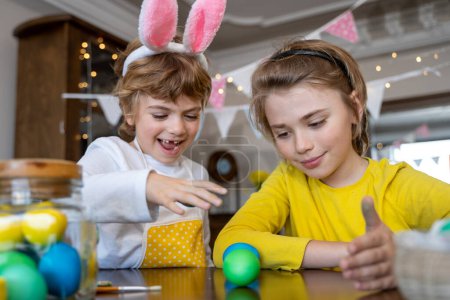 Foto de Easter Family traditions. Two caucasian happy children with bunny ears dye and decorate eggs with paints for holidays playing together. Kids having fun. - Imagen libre de derechos