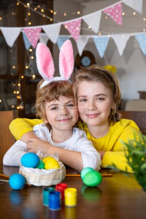 Photo for Easter Family traditions. Two caucasian happy children with bunny ears dye and decorate eggs with paints for holidays while sitting together at home table. Kids embrace and smile in cozy. Vertical - Royalty Free Image