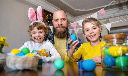 Photo for Easter Family traditions. Father and two caucasian happy children with bunny ears dye and decorate eggs with paints for holidays while sitting together at home table. Looking at camera - Royalty Free Image