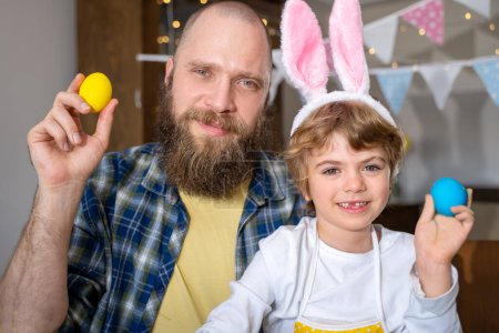 Foto de Easter Family traditions. Father and caucasian happy child with bunny ears dye and decorate eggs with paints for holidays while sitting together at home table. Vertical - Imagen libre de derechos