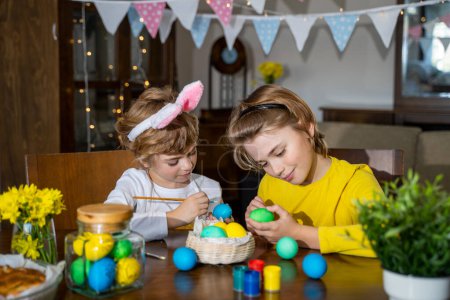 Photo for Easter Family traditions. Two caucasian happy children with bunny ears dye and decorate eggs with paints for holidays while sitting together at home table. Kids having fun together - Royalty Free Image