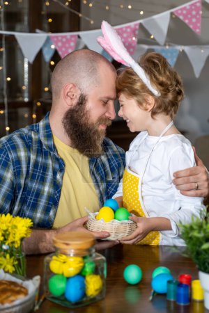 Téléchargez les photos : Traditions familiales de Pâques. Père et caucasien heureux enfant avec des oreilles de lapin teindre et décorer les œufs avec des peintures pour les vacances tout en embrassant ensemble à la table à la maison. Vertical - en image libre de droit