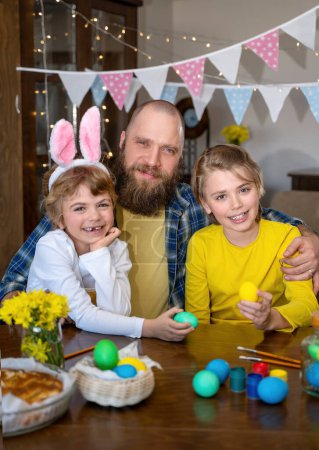 Foto de Easter Family traditions. Father and two caucasian happy children with bunny ears dye and decorate eggs with paints for holidays while sitting together at home table. Kids embrace and smile in cozy. - Imagen libre de derechos