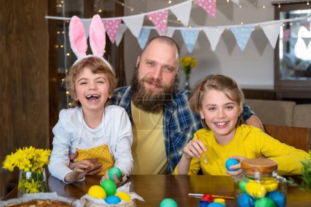 Téléchargez les photos : Easter Family traditions. Father and two caucasian happy children with bunny ears dye and decorate eggs with paints for holidays while sitting together at home table. Kids embrace and smile in cozy - en image libre de droit