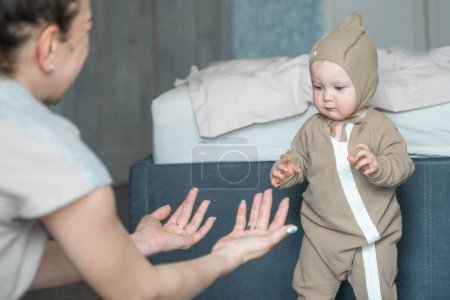 Photo for Little toddler infant in beige bodysuit and cap learning to walk first steps on home carpet hold loving mother hands. Small caucasian baby child learn walking with mom support care. Childcare concept. - Royalty Free Image