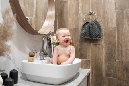 Photo for Baby bathing in bathroom sink. Child playing with water in cozy bathroom. Little toddler take shower. Water fun for kids. Hygiene and skin care for children. Bath room interior - Royalty Free Image