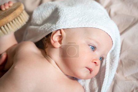 Photo for Baby in bath towel after hygiene procedures, lying on parent bed in bedroom. Hold comb Hygiene and skin care for children. - Royalty Free Image