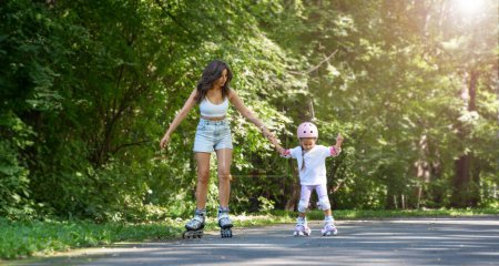 Téléchargez les photos : Mère enseignant à sa fille à patiner sur des patins à roues alignées dans un parc public en été. Loisirs en famille jeu d'activité sportive de plein air. Copier l'espace négatif - en image libre de droit